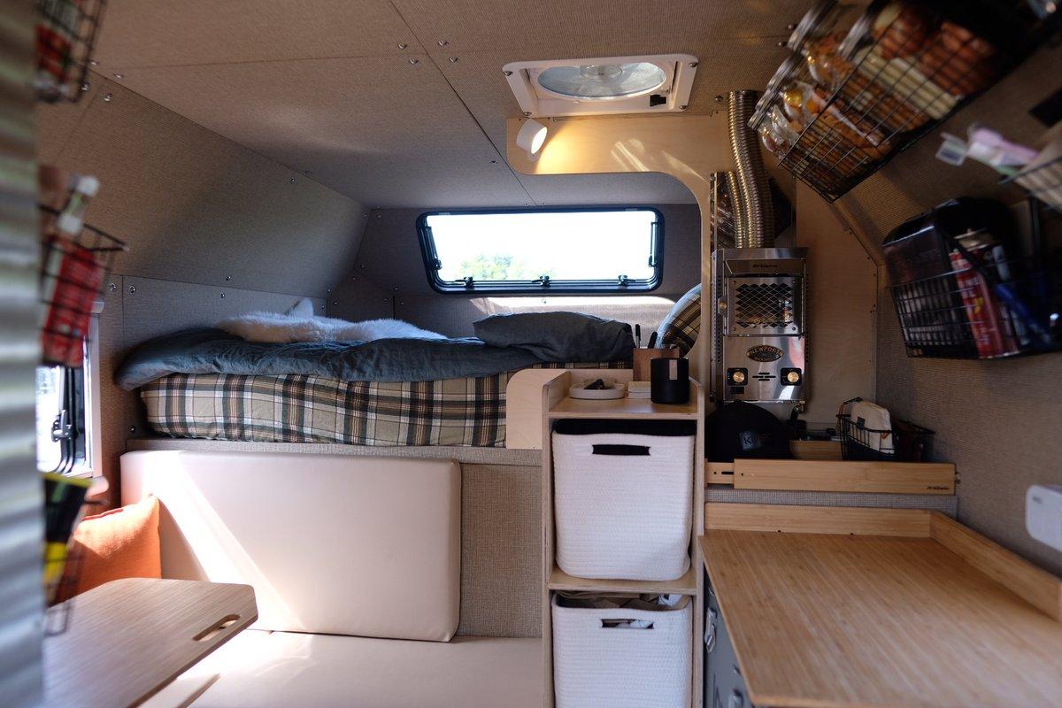 Wide interior view of a Kimbo 8 hard-side aluminum truck bed camper from the entry doorway — the cabover bed at upper left, a U-shaped dinette with an orange pillow at lower left, the bamboo galley counter at center-right, and the Dickinson Newport heater venting through a stainless chimney along the back wall.