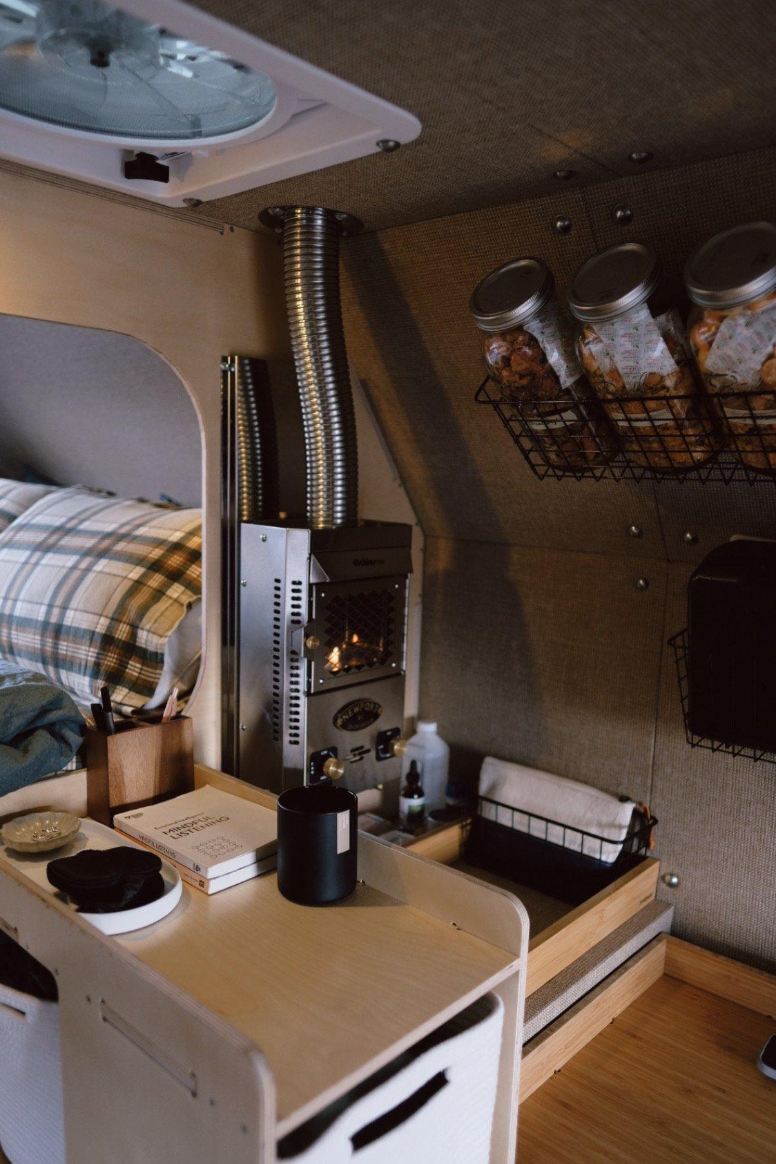 Vertical detail of the stainless Dickinson Newport propane heater inside a Kimbo 8 hard-side aluminum truck bed camper, with the corrugated stainless chimney climbing past a MaxxAir roof vent, three sealed mason jars in a wire pantry basket at upper right, and a book titled Mindful Listening on the bedside counter.