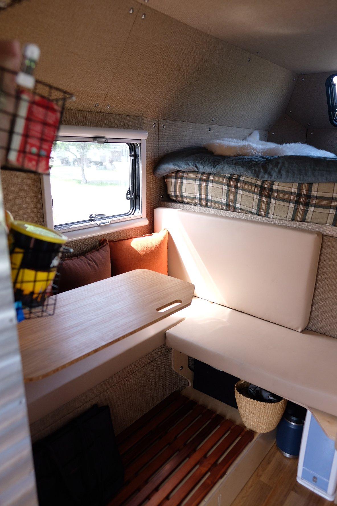 Kimbo 8 hard-side aluminum truck bed camper U-shaped dinette with two orange and one brown throw pillow, the bamboo swivel table folded down, an Arctic Tern side window looking out to a green park, the cabover bed visible above with a plaid duvet.