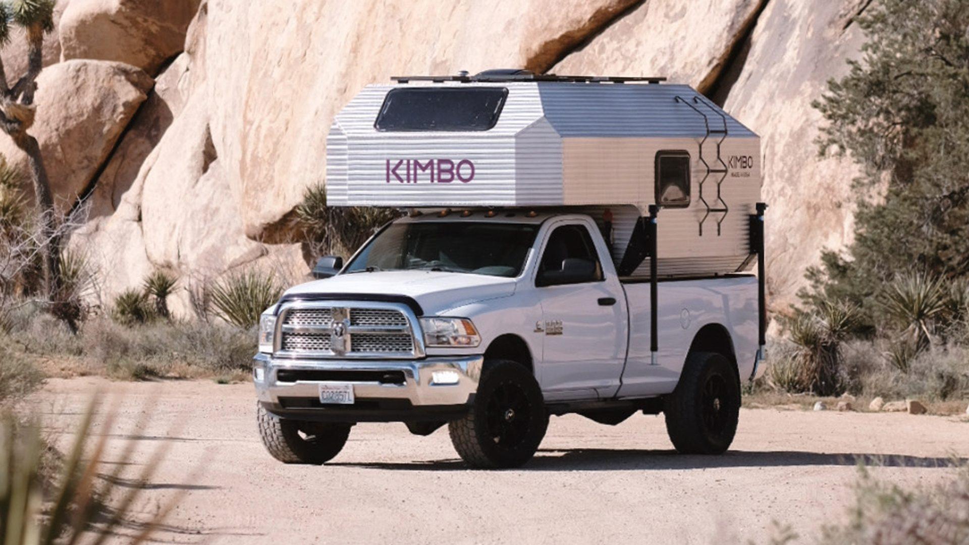 White Dodge Ram 2500 with the Kimbo 8 hard-side aluminum truck bed camper boondocking among large granite boulders at Joshua Tree National Park, side-quarter view at golden hour.
