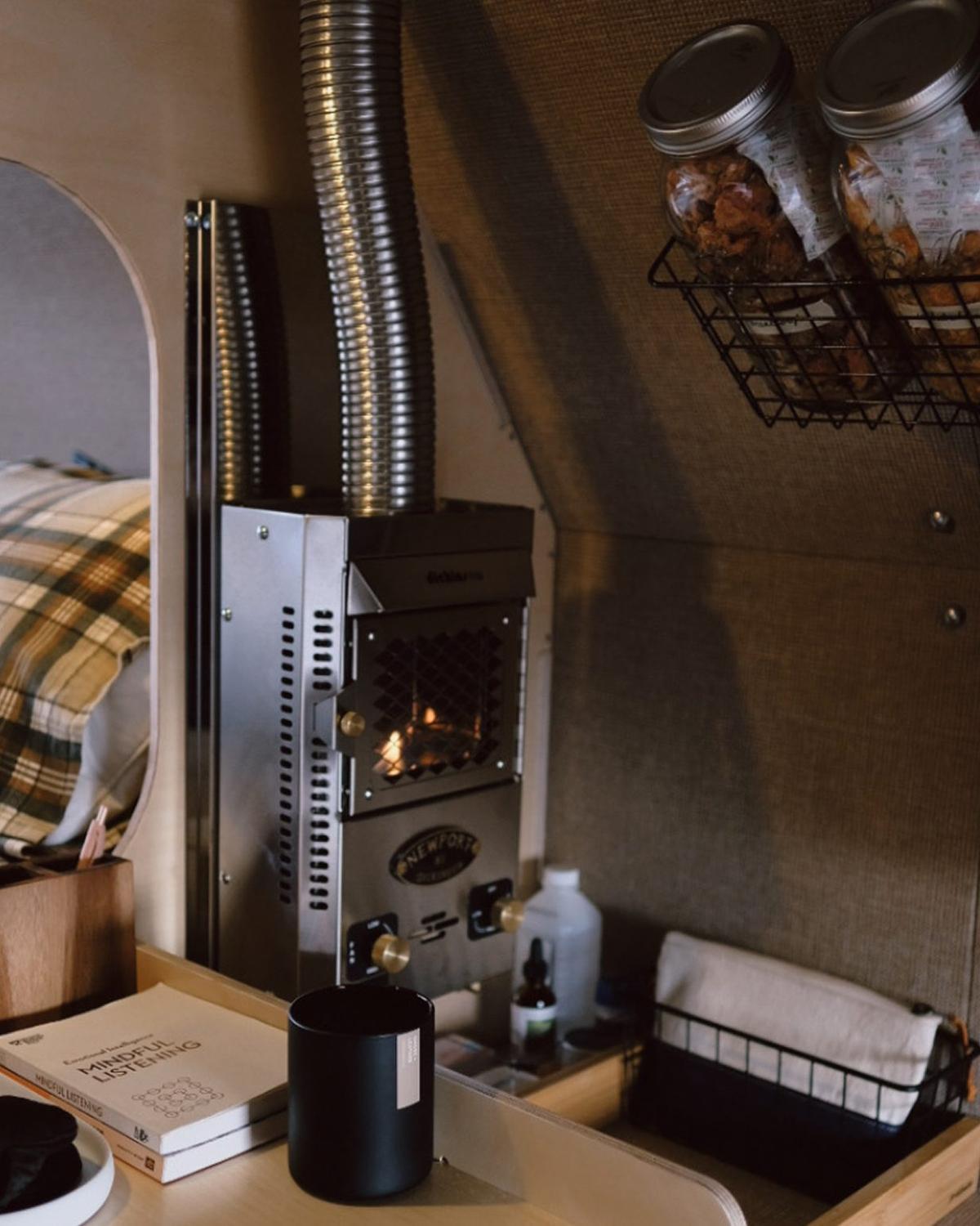 Interior of a Kimbo 8 hard-side aluminum truck bed camper showing the stainless Dickinson Newport propane heater with its corrugated stainless chimney climbing to the ceiling, a wire-basket pantry of sealed mason jars on the right wall, a plaid pillow in the cabover bed and a book titled Mindful Listening on the bedside counter.