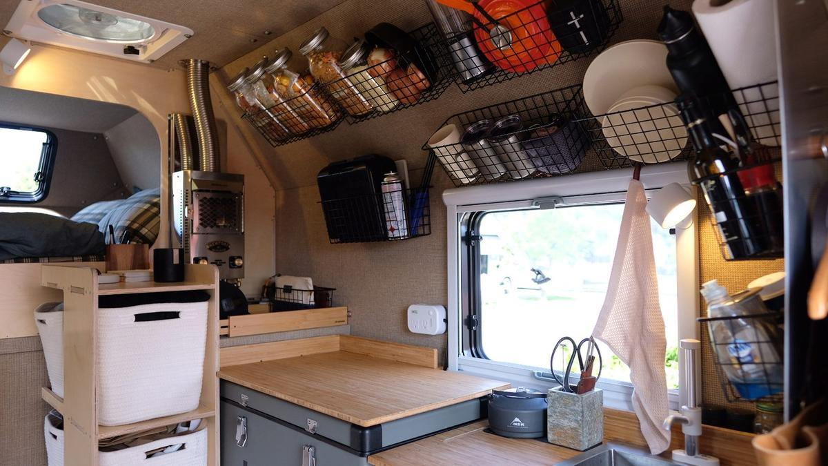 Wide-angle Kimbo 8 galley interior with a deep stainless sink, bamboo prep counter, ICECO 12V fridge, a full wall of mounted wire baskets stocked with kitchen tools, and an Arctic Tern double-pane window looking out toward the dinette.