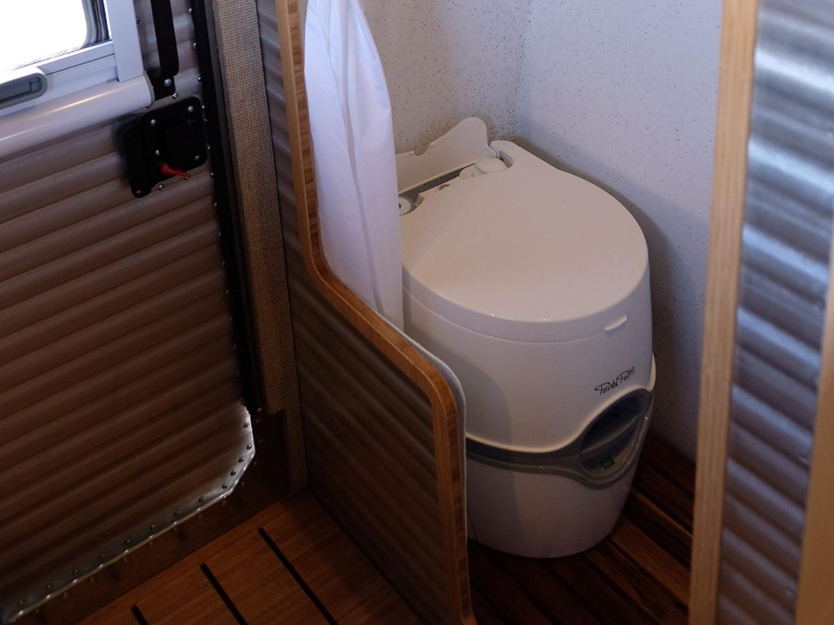 Dedicated wet bath inside a Kimbo 8 hard-side aluminum truck bed camper — fully enclosed cassette toilet behind a sliding shower curtain, drained teak floor slats, corrugated aluminum walls, and a window-side mudroom wall.