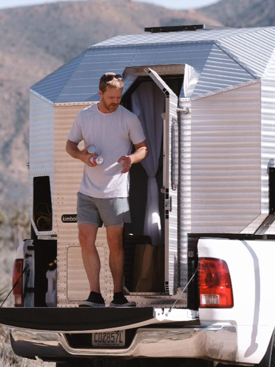 Kimbo 8 owner standing on the open tailgate of his white Dodge Ram 2500 truck with the Kimbo 8 hard-side aluminum truck bed camper installed in the bed, holding two beverage cans, photographed from low rear-three-quarter at a Joshua Tree desert pull-off.