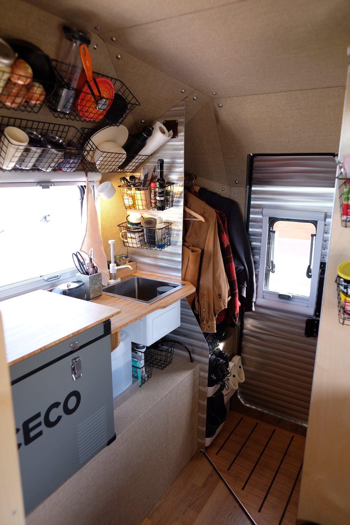 Looking through the entry doorway of a Kimbo 8 hard-side aluminum truck bed camper from outside — bamboo coat rod with red flannel and tan jackets, mounted wire-basket pantry above the stainless sink and induction cooktop, ICECO 12V refrigerator at lower left, hiking boots tucked under the entry step.