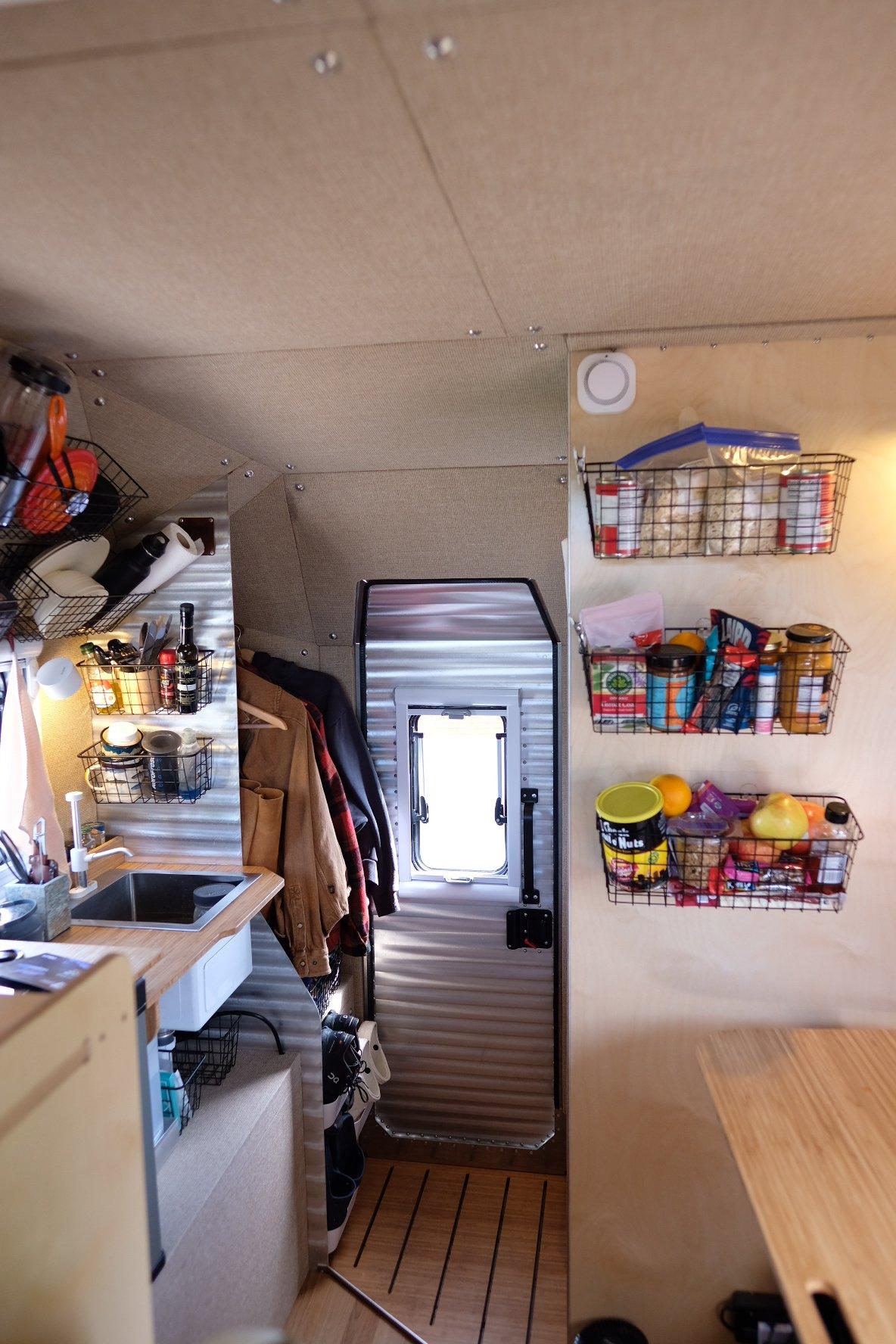 Looking into a Kimbo 8 hard-side aluminum truck bed camper through the open rear door, framed by the corrugated aluminum entry wall — three mounted wire-basket pantry organizers full of food on the right wall, the bamboo galley counter with stainless sink on the left wall, a smoke / propane alarm on the ceiling above.