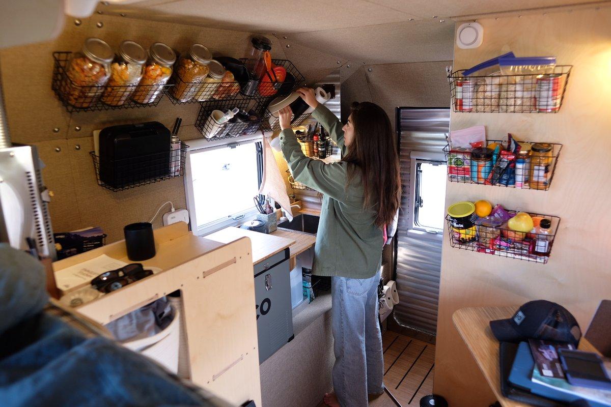 Female Kimbo 8 owner stretching to reach a wax-paper roll from the upper wire-basket pantry inside a Kimbo 8 hard-side aluminum truck bed camper, full-wall mason-jar pantry behind, the bamboo galley counter at left with the ICECO 12V fridge and a black mug.