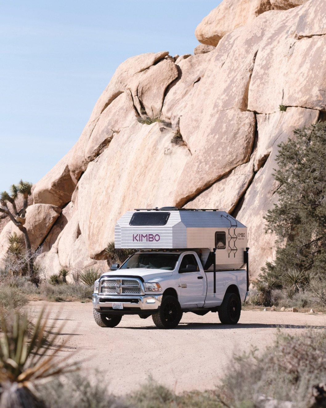 White Dodge Ram 2500 with the Kimbo 8 hard-side aluminum truck bed camper installed, photographed front-three-quarter on a dirt pull-off in Joshua Tree National Park, a single massive granite boulder rising behind the truck and a foreground Joshua tree at left.