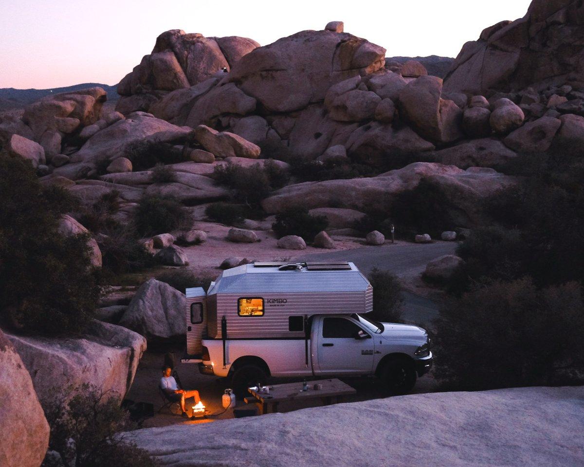 White Dodge Ram 2500 with the Kimbo 8 hard-side aluminum truck bed camper parked against a granite boulder formation in Joshua Tree National Park at dusk, the owner sitting in a camp chair beside a small campfire and propane lantern, the cabover window glowing warm orange from the interior lights against a pink and lavender desert sky.