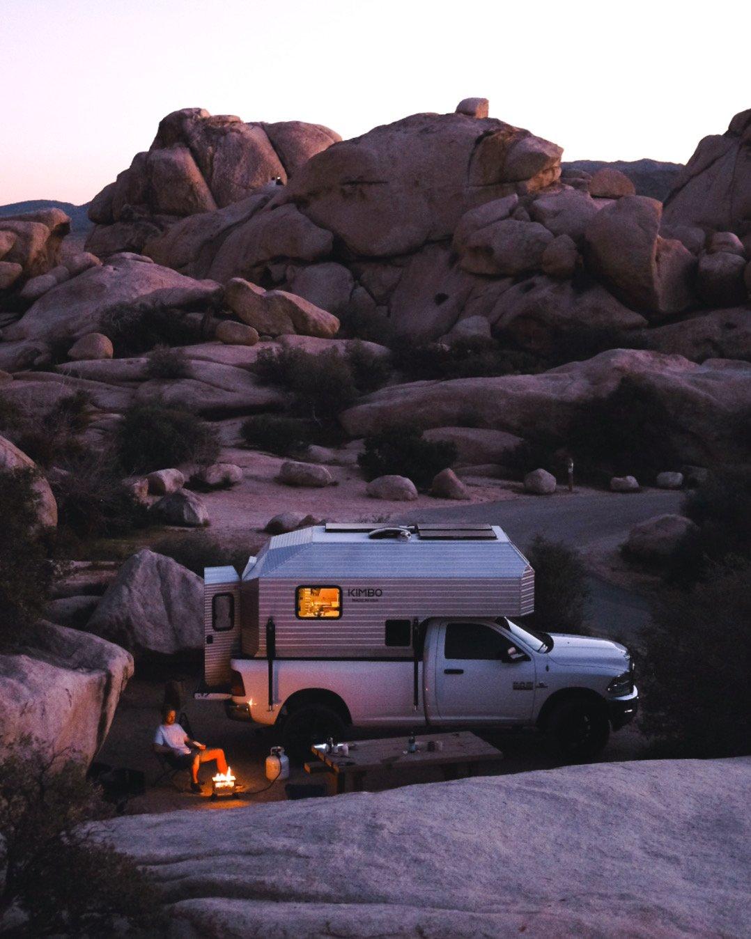 Vertical view of a white Dodge Ram 2500 with the Kimbo 8 hard-side aluminum truck bed camper parked beneath a towering granite boulder formation in Joshua Tree National Park at last light, the owner sitting beside a small campfire with the cabover window glowing warm against the dusk sky.