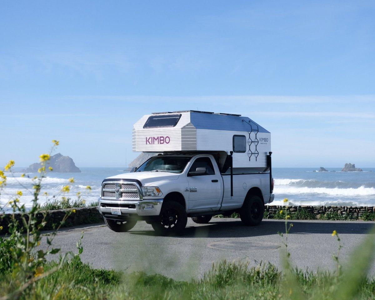 White Dodge Ram 2500 with the Kimbo 8 hand-riveted aluminum truck bed camper installed, photographed front-three-quarter at a coastal pull-off on the Oregon coast, breaking waves and sea stacks behind, wildflowers in the foreground.