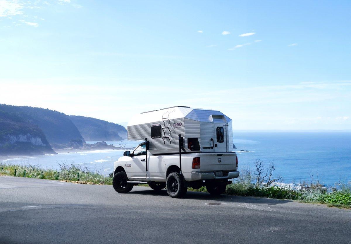 White Dodge Ram 2500 with the Kimbo 8 hard-side aluminum truck bed camper parked at a Pacific coastal overlook, photographed rear-three-quarter from the road shoulder, the side ladder folded against the camper, the coastline curving away behind down a long beach.