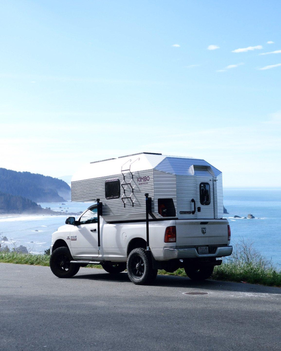 Vertical view of a white Dodge Ram 2500 with the Kimbo 8 hard-side aluminum truck bed camper parked at a Pacific coastal overlook in clear weather, photographed rear-quarter from below the road, the breaking surf and coastal headlands stretching to the horizon.