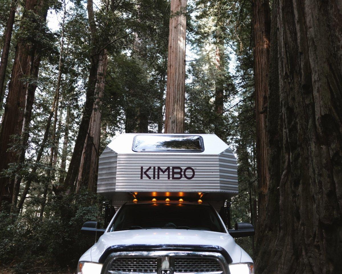 Looking up from below the front grille of a white Dodge Ram 2500 with the Kimbo 8 hard-side aluminum truck bed camper installed, the towering coastal redwoods stretching impossibly tall into the canopy directly above and the KIMBO wordmark prominent on the front of the cabover.