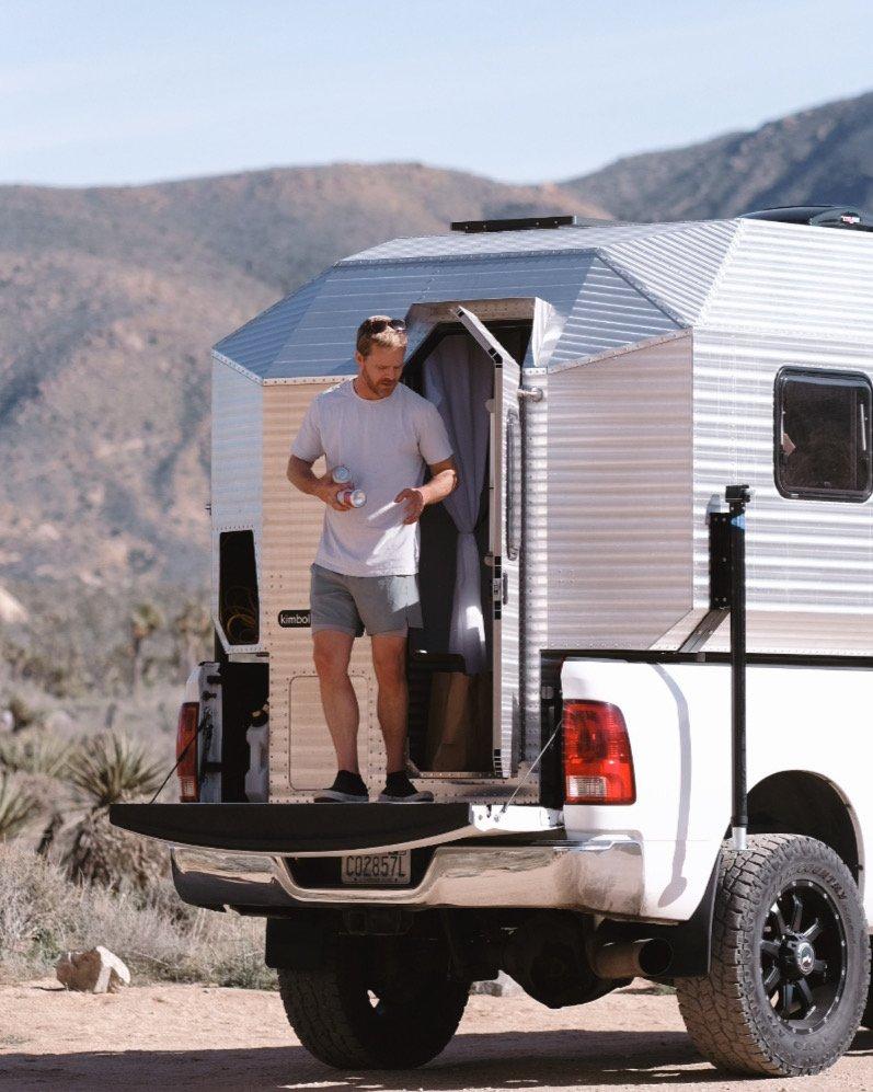Wider rear-three-quarter view of a Kimbo 8 hard-side aluminum truck bed camper installed on a white Dodge Ram 2500 truck in the high Mojave Desert north of Joshua Tree, the owner stepping out of the open rear door onto the tailgate holding two beverage cans, dry creosote and distant mountains in the background.