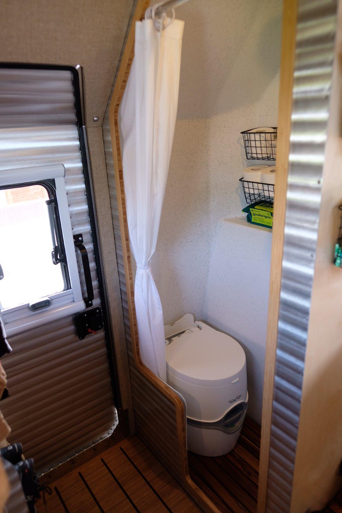 Interior portrait detail of the wet bath inside a Kimbo 8 hard-side aluminum truck bed camper — Thetford cassette toilet with a soft-fold shower curtain pulled half-back, toilet paper stored in a mounted wire basket above, teak floor slats below, corrugated aluminum walls, and a small Arctic Tern window with insulated blackout shade at left.