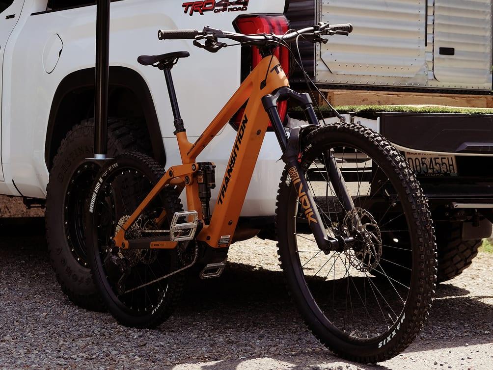 An orange Transition Bikes mountain bike loaded on the rear hitch rack of a white Toyota Tacoma TRD Off-Road.